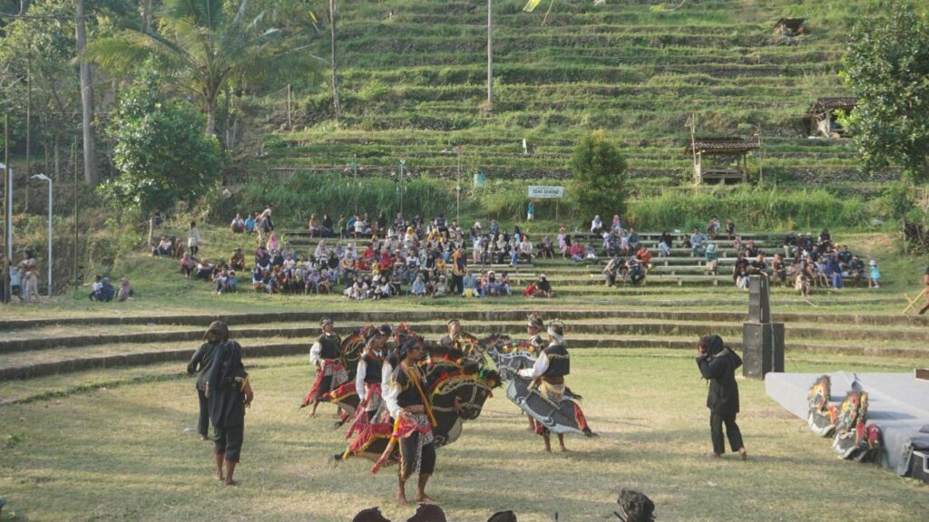 Dancers in colorful traditional costumes perform on a grassy field as a large audience sits on terraced benches along a hillside